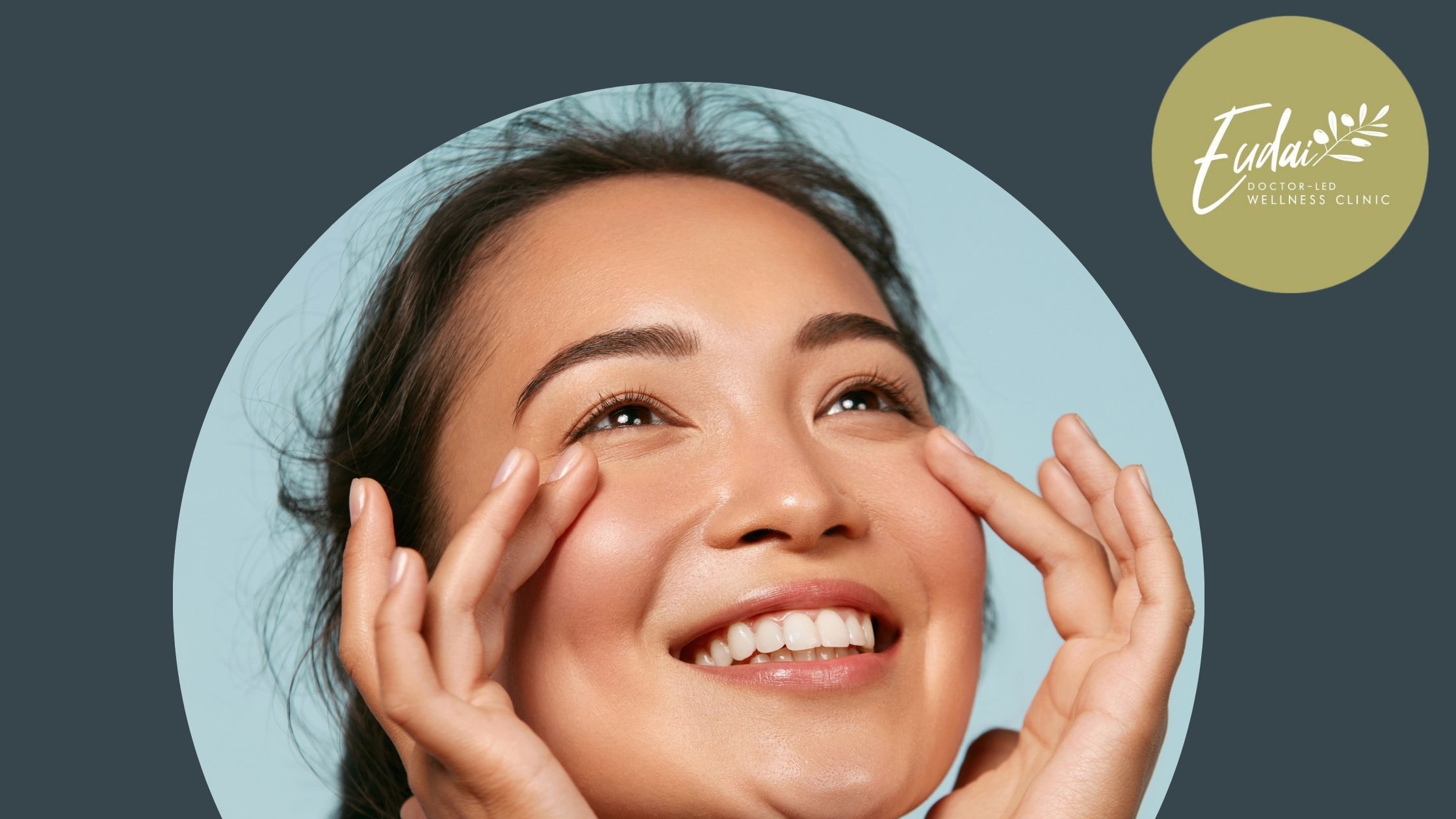 A joyful woman with radiant skin lightly pinching her cheeks, showcasing a youthful complexion, indicative of the quality care at Eudai Wellness Clinic.