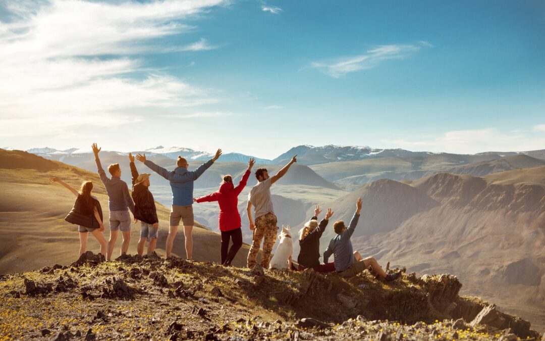 A group of people stands on top of a mountain, facing the expansive landscape. They raise their arms in celebration, with snowy peaks and valleys stretching out beneath a blue sky.