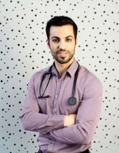 A man, Dr. Dimi, wearing a light purple patterned shirt with a stethoscope around his neck. He is standing with arms crossed in front of a light background with black dots.