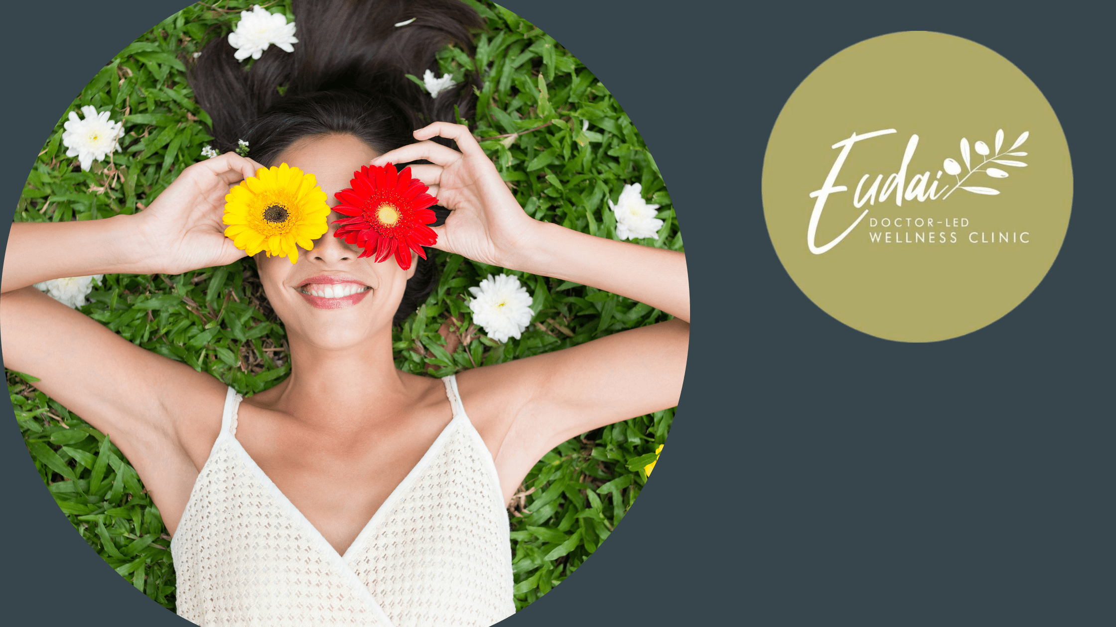 A playful image of a woman lying on the grass, holding a red and yellow flower over her eyes. Her hair is spread around her head, and there are white flowers scattered on the grass, creating a whimsical, fun atmosphere.