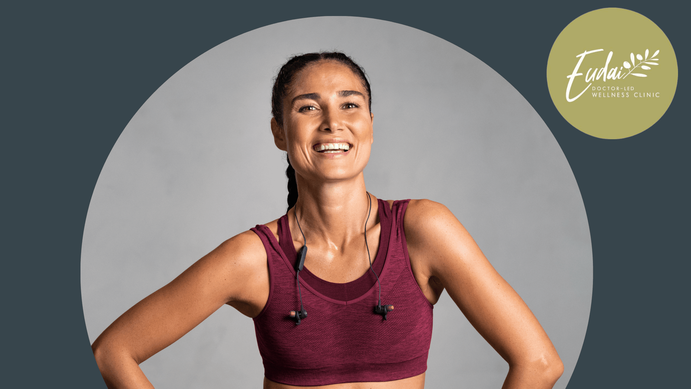 A woman with braided dark hair stands confidently in a maroon sports bra. She is smiling, looking upward, and has earphones around her neck. She appears to have finished a workout and is radiating positivity.