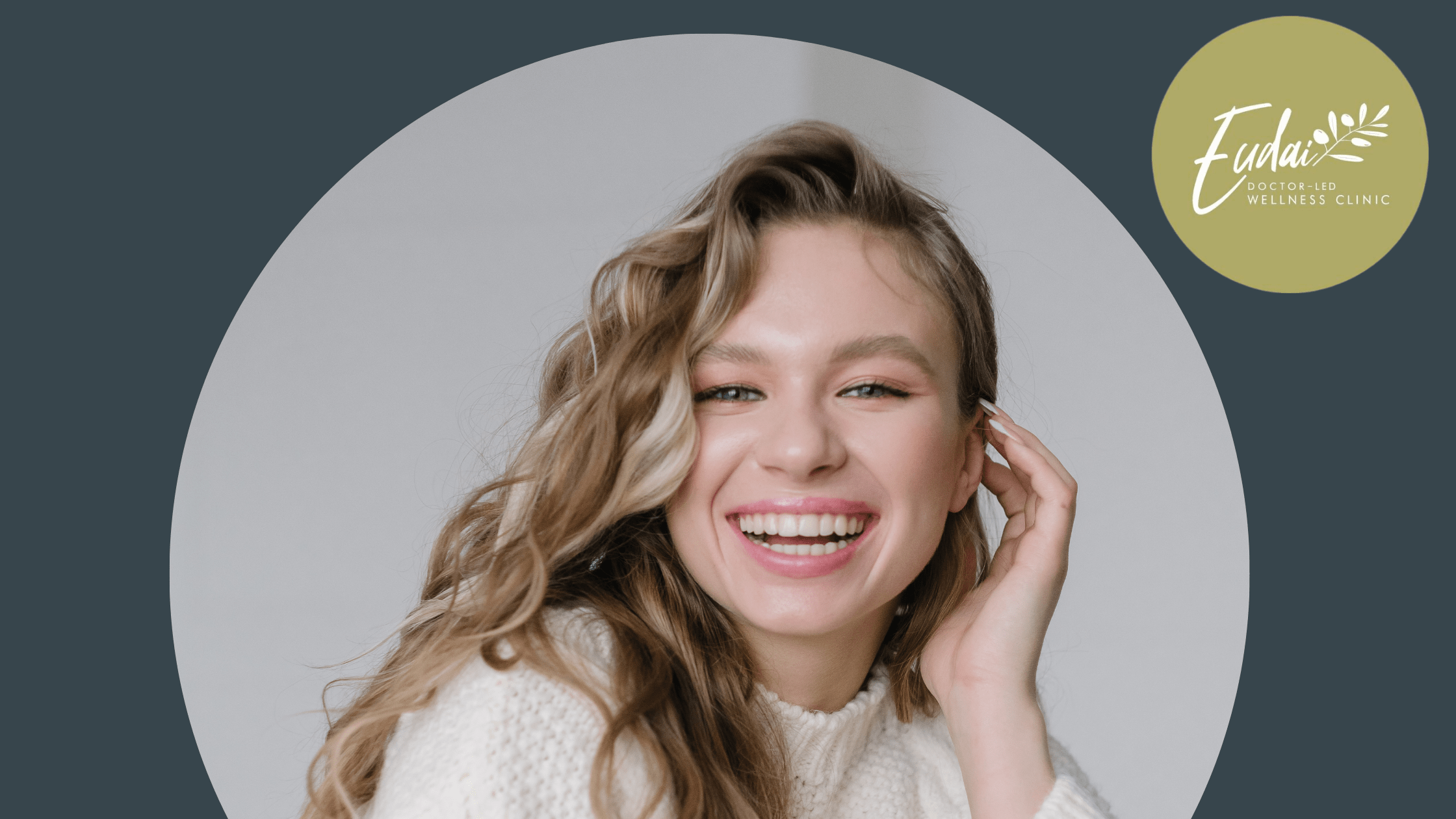 A young woman with long, wavy blonde hair is smiling brightly. She is wearing a cozy white sweater and casually playing with her hair. Her expression conveys joy and energy, with a simple background enhancing her look.