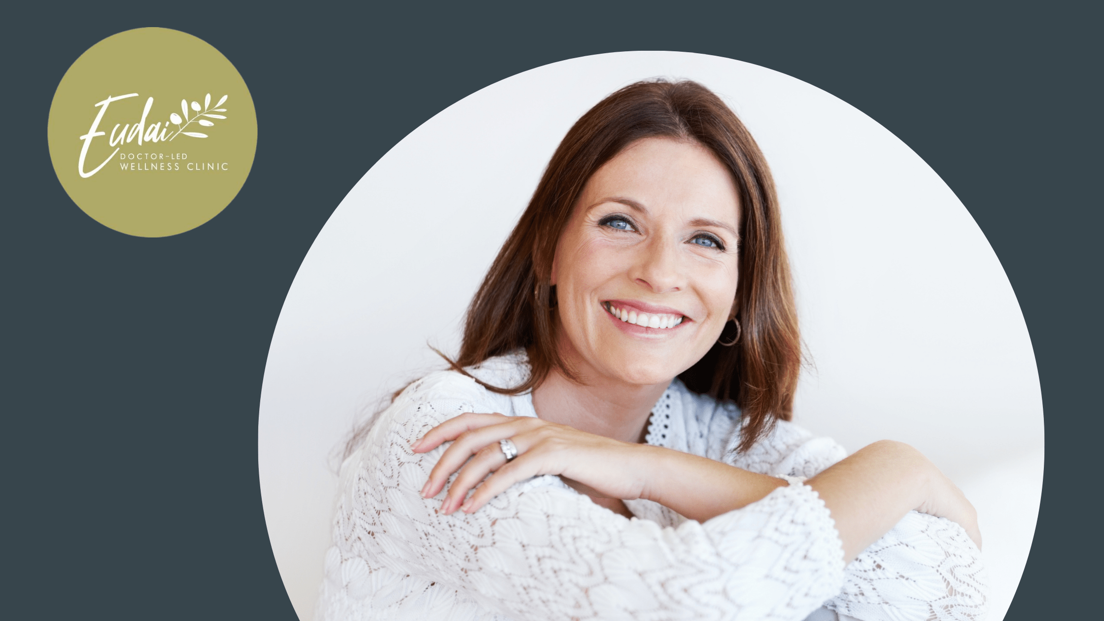 A woman with long brown hair, smiling warmly while wearing a white, long-sleeved top. She has her arms crossed on her lap and is seated in a bright, minimalist room. The Eudai Clinic logo is visible in the top left corner on a dark gray background.