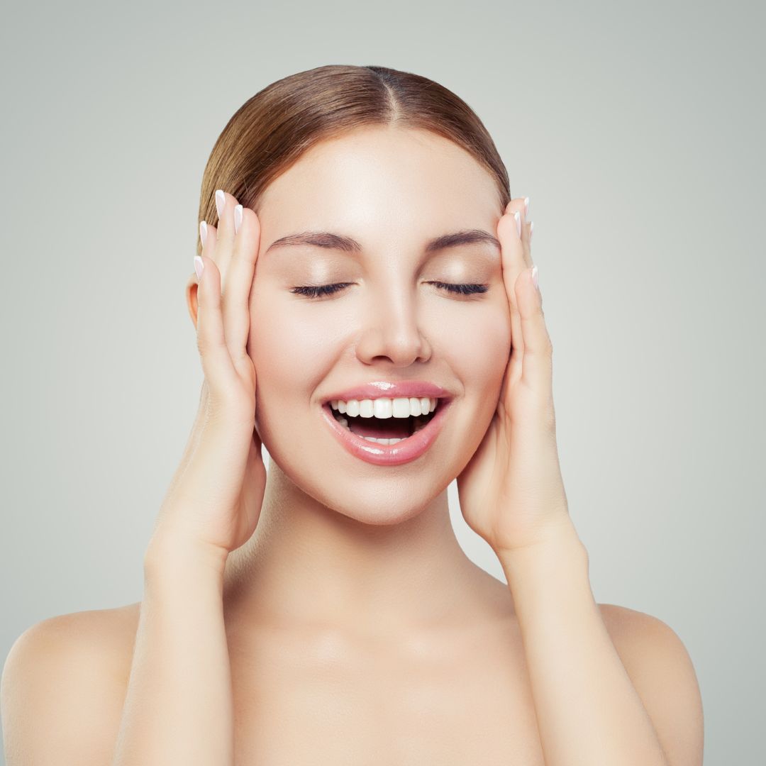 Close-up portrait of a young woman with clear skin smiling with her eyes closed, gently holding her face with both hands against a neutral background.
